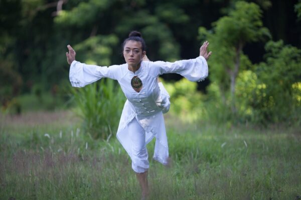 A woman in all white oriental outfit doing crane qigong outside